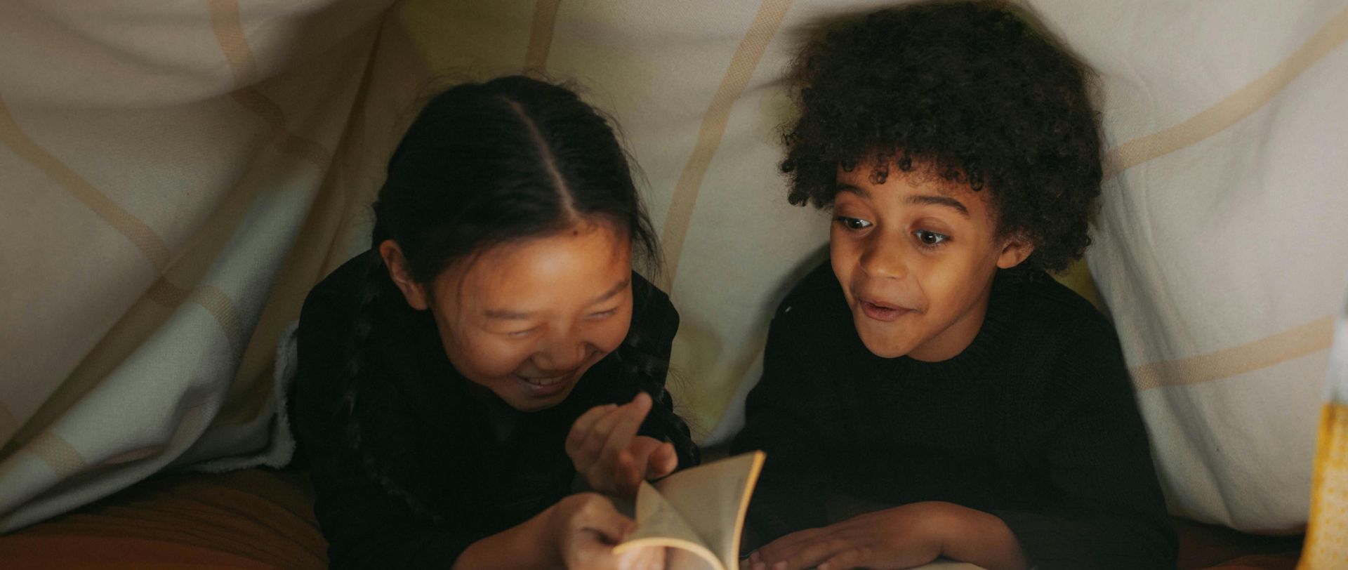 Two children read a book together under a cozy blanket fort, illuminated by soft lighting.